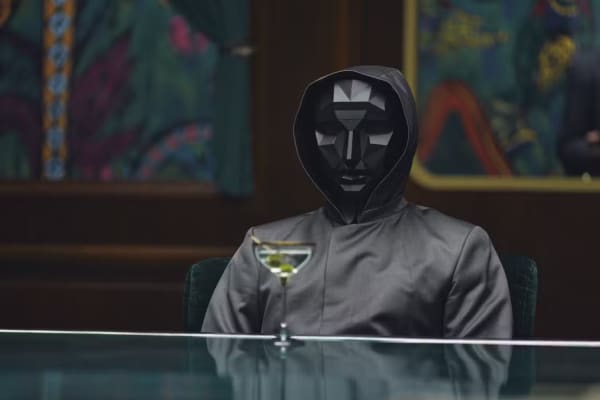 A person in grey mask and grey hoodie sits in front of a cocktail.