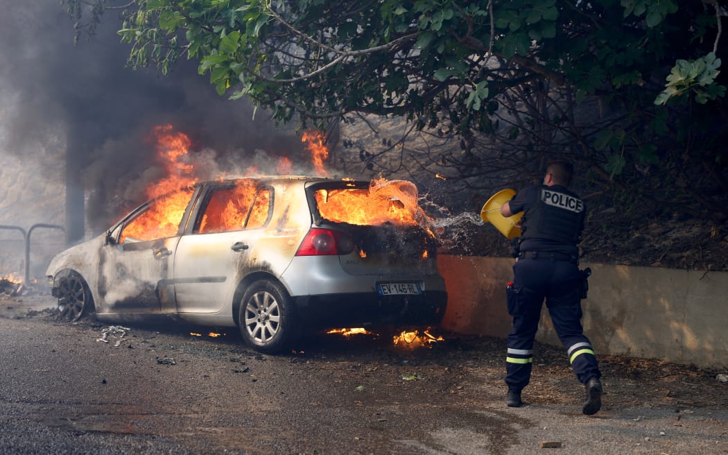 A police officer tries to put off the fire in a car during a wildfire, in L'Estaque district of Marseille, southern France on July 8, 2025. The fire that started on July 8, 2025 in Les Pennes-Mirabeau (Bouches-du-Rhone), a town bordering Marseille, has entered France's second-largest city, where some residents were urged to shelter in place, according to a warning message at 4:00 PM from the prefect of Bouches-du-Rhone. (Photo by Clement MAHOUDEAU / AFP)