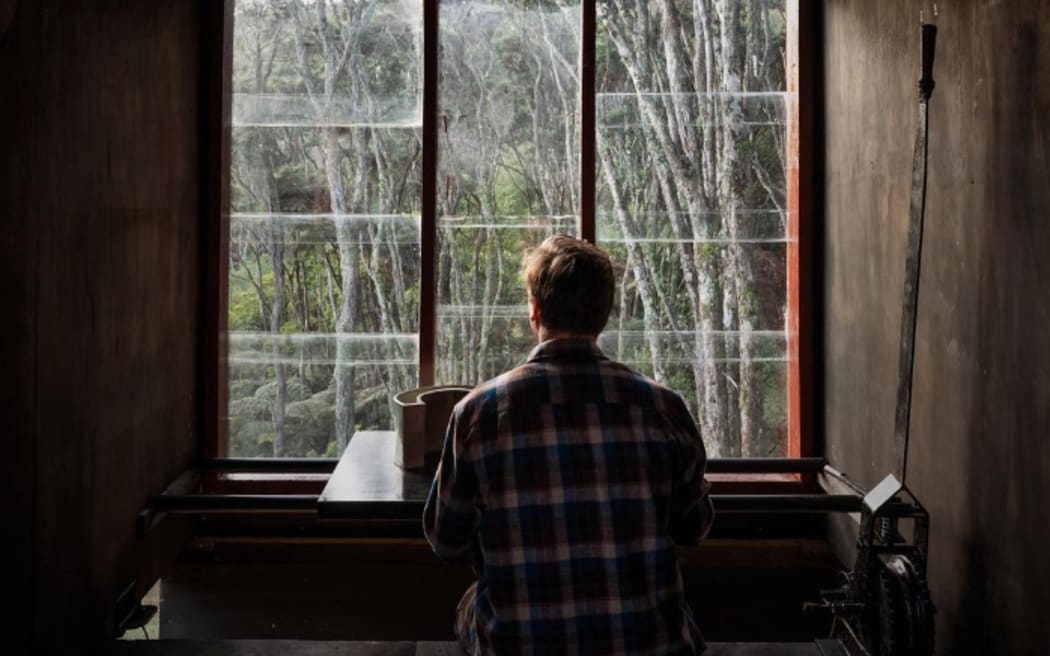 Man sits in front of a window in a darkened room, his back to camera.