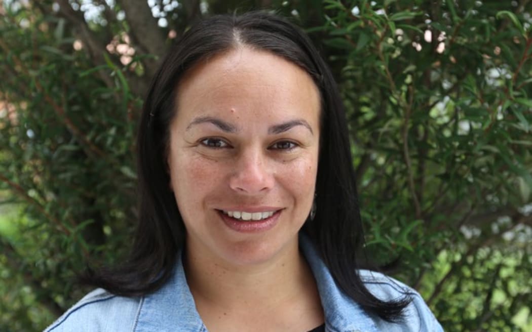 A woman wearing a black shirt and denim jacket smiles at the camera. She has long dark brown hair and is standing in front of a tree.