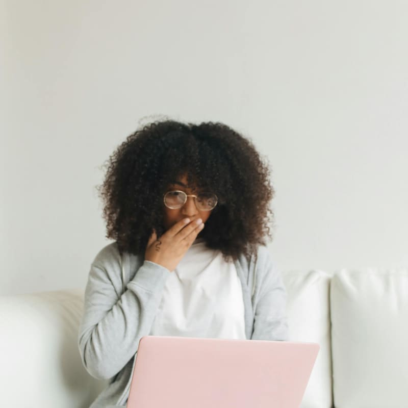A woman wearing a white tshirt and grey cardigan covers her mouth with her hand in surprise as she looks at a laptop.ggen