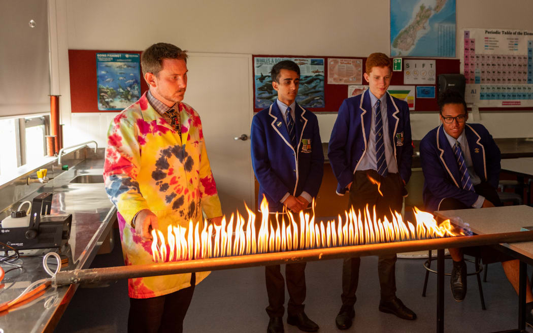 A man in a tie-dyed lab coat stands next to two high school boys in blazers behind a pipe emitting a row of flames.
