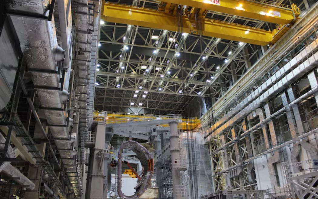 An indoor multi-storey hall filled with steel beams and construction activity, including a large D-shaped segment suspended at the far end of the space.