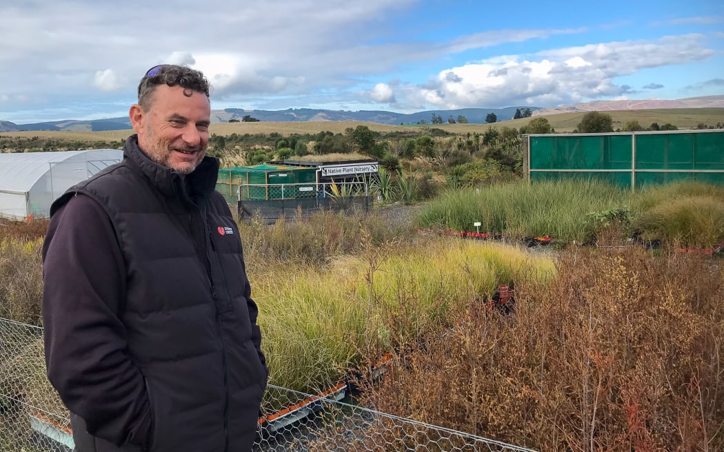 A man wearing a puffer vest and long-sleeved dark jumper stands in front of rows of native grass seedlings in a nursery. He has his hands in his pockets and is smiling at the camera. Buildings and greenhouses are visible in the background. The sky is cloudy with patches of blue.