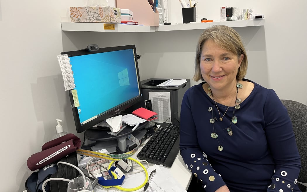 A woman sits at her desk in a medical office. The desk is strewn with papers, equipment, with a computer desktop sitting in the centre. The woman is facing the camera in her chair, smiling.