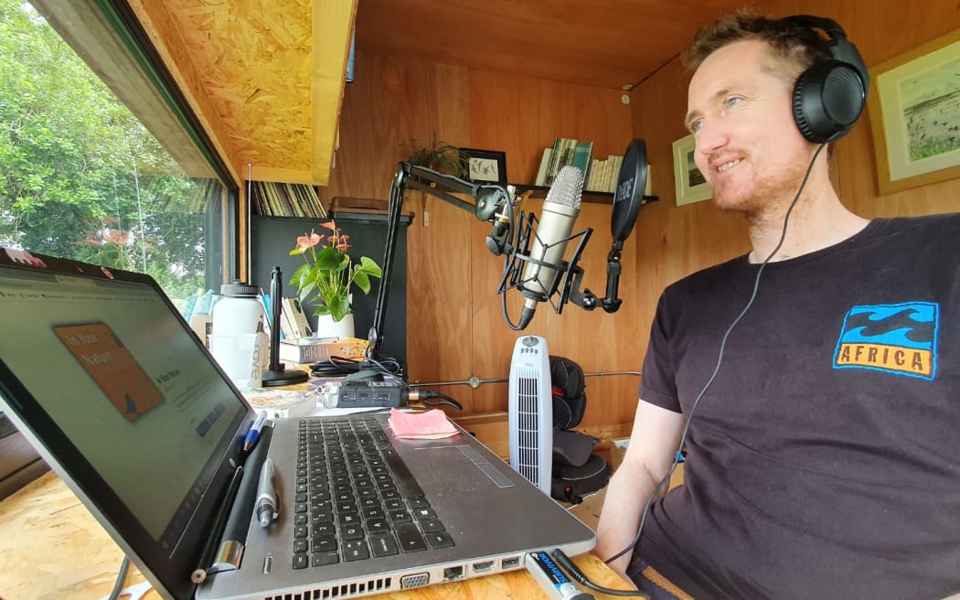 A man wearing headphones and a tshirt sits at a microphone in front of a laptop.