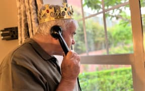 Tom, an elderly man in a brown t-shirt, looks out a window wearing a golden crown and holding an old-fashioned black rotary phone.