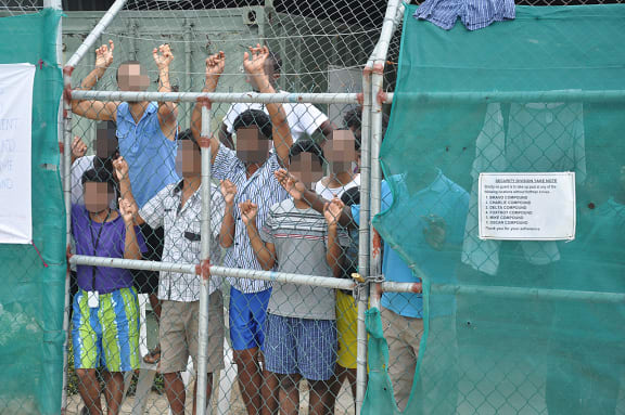 Asylum seekers behind behind a fence on which a sign orders guards to carry the hooked knives used to cut the rope of people who attempt to hang themselves.