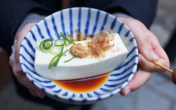 A blue striped bowl containing a piece of tofu bathed in soy sauce with spring onion garnish