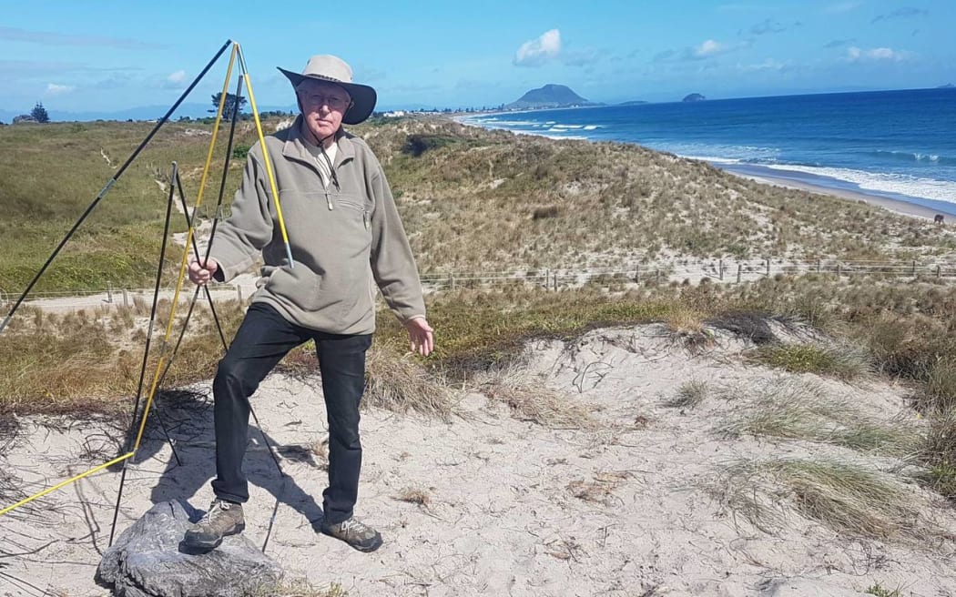 Pāpāmoa Beach resident Bruce Hutchinson stands in a bare patch of damaged and trampled dunes and holds some of the rubbish left behind following the party on 26 November, 2022.