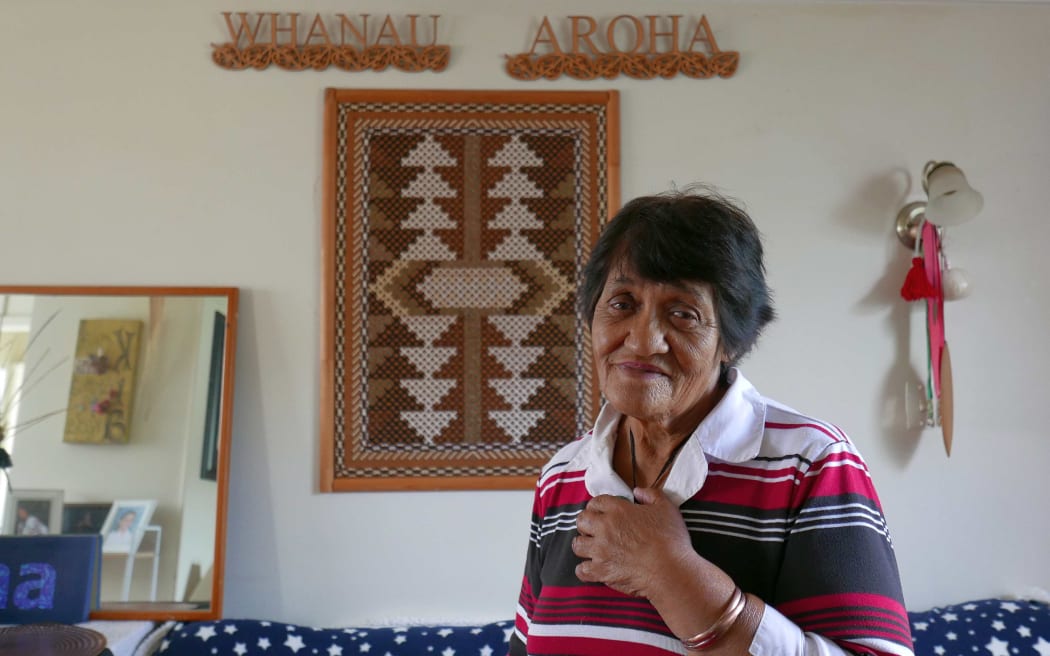 Older woman at home with  carving behind saying "whanau" and "aroha"
