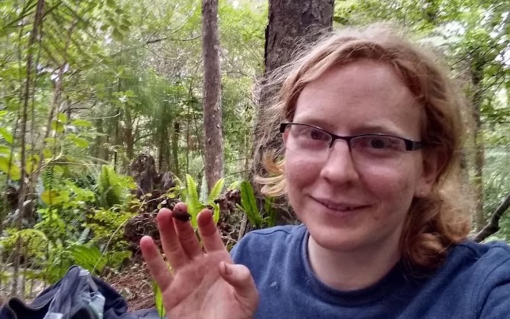 Luna is in the bush, wearing glasses, holding up her right hand which has a beetle on one of her fingers.