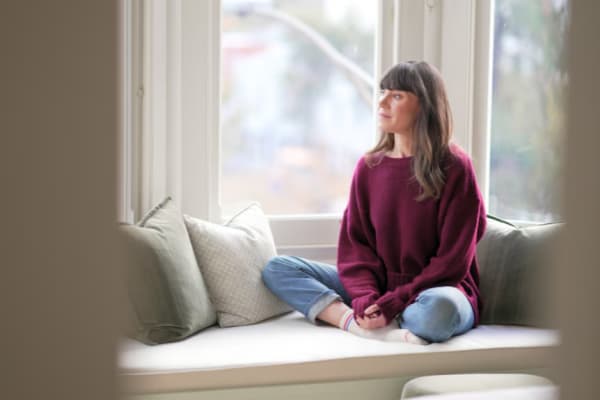 A woman in a berry-coloured sweater and jeans sits cross-legged by a window, looking out.