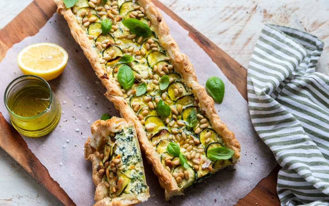 A rectangular shaped spinach tart decorated with pine nuts and basil leaves, on a wooden board lined with baking paper.