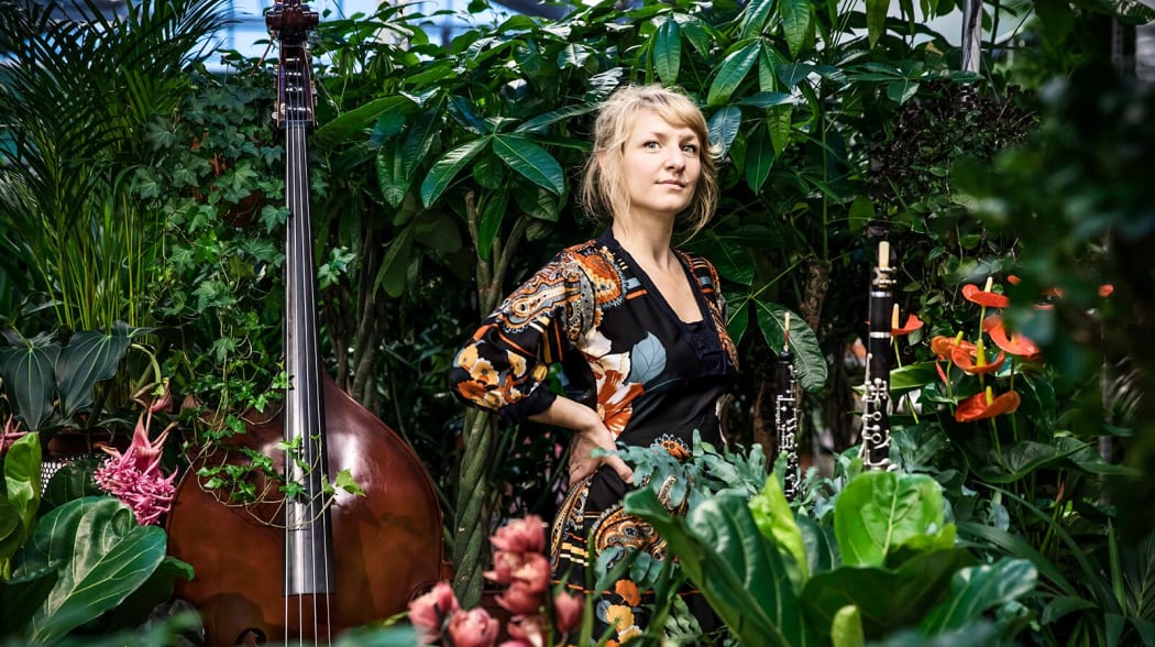 Promo shot of Swedish composer Andrea Tarrodi. She stands surrounded by greenery and musical instruments
