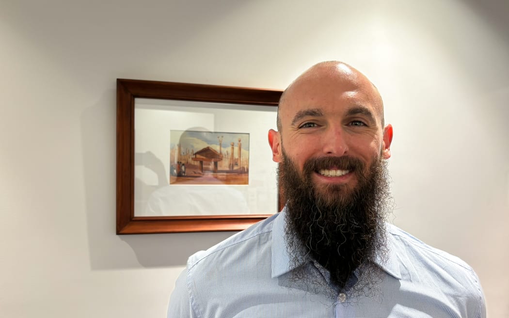 A mid-shot of a man wearing a light blue shirt with a long dark beard and no hair smiles widely at the camera. He is standing in front of a small painting of a Māori storehouse in a wooden frame.