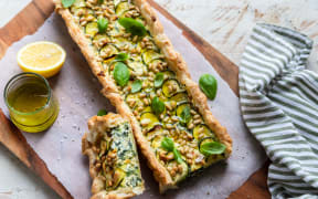 A rectangular shaped spinach tart decorated with pine nuts and basil leaves, on a wooden board lined with baking paper.