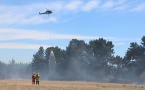 Firefighters watch on as a helicopter dumps water on a wildfire near Kirwee, Canterbury. February 5, 2024.