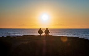 A couple sit together on fold out chairs watching a sunset over the sea.