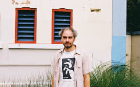 Peter McCall stands in front of a building wearing a beige short sleeved shirt over a white t=shirt with a black graphic print.