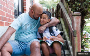 A stock photo of a father and son sitting on the steps outside a house, smiling. The father embraces the son.