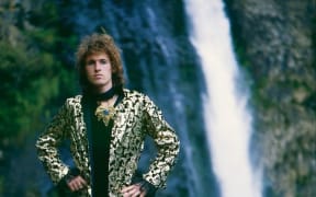 An image of New Zealand musician Andrew Fagan pictured in front of a waterfall. Andrew has shaggy brown hair and is wearing a shiny black and gold suit, with a black shirt and elaborate gold corsage.