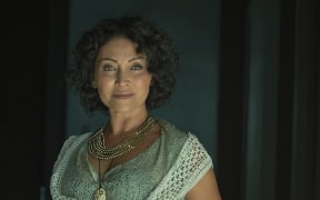Tina Cross smiles slightly at the camera while clutching a knitted vest over a green dress.