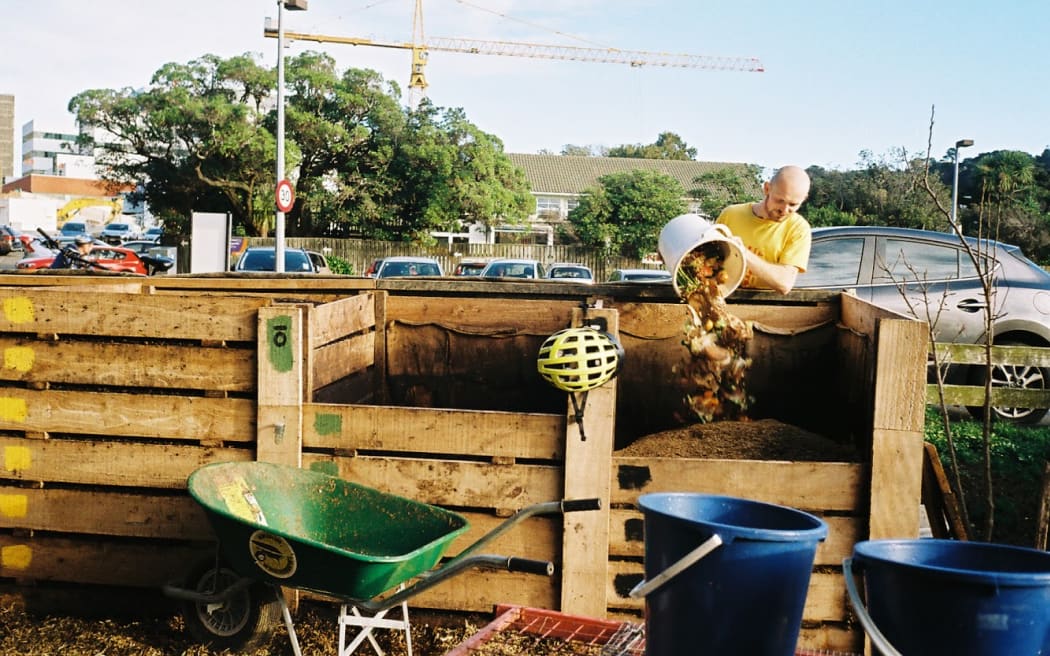 A man deposits compost from a white bucking it a composting pile, which is located inside a wooden box.