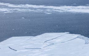 A wide shot of ice floes breaking apart, as seen from a high angle. Beyond the ice edge, there is open dark water, then a line of mushy ice.