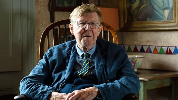Alan Bennett is a senior man with combed brown hair, glasses and a tie, sitting in a wooden chair.