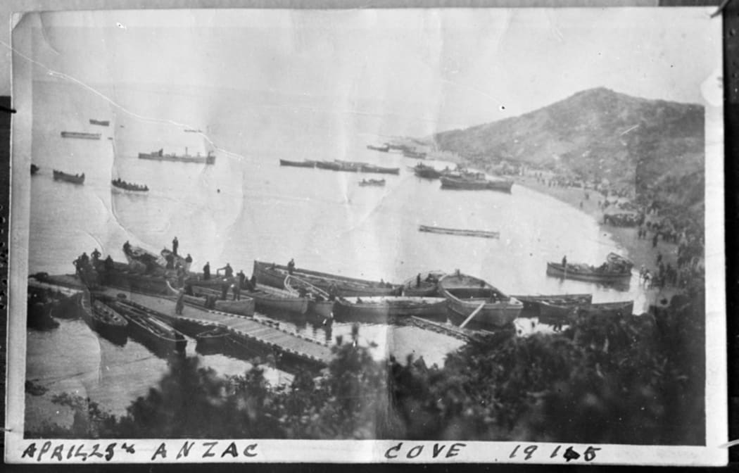 Landing craft moored at a pontoon with other small vessels in the background and soldiers gathered on the beach.