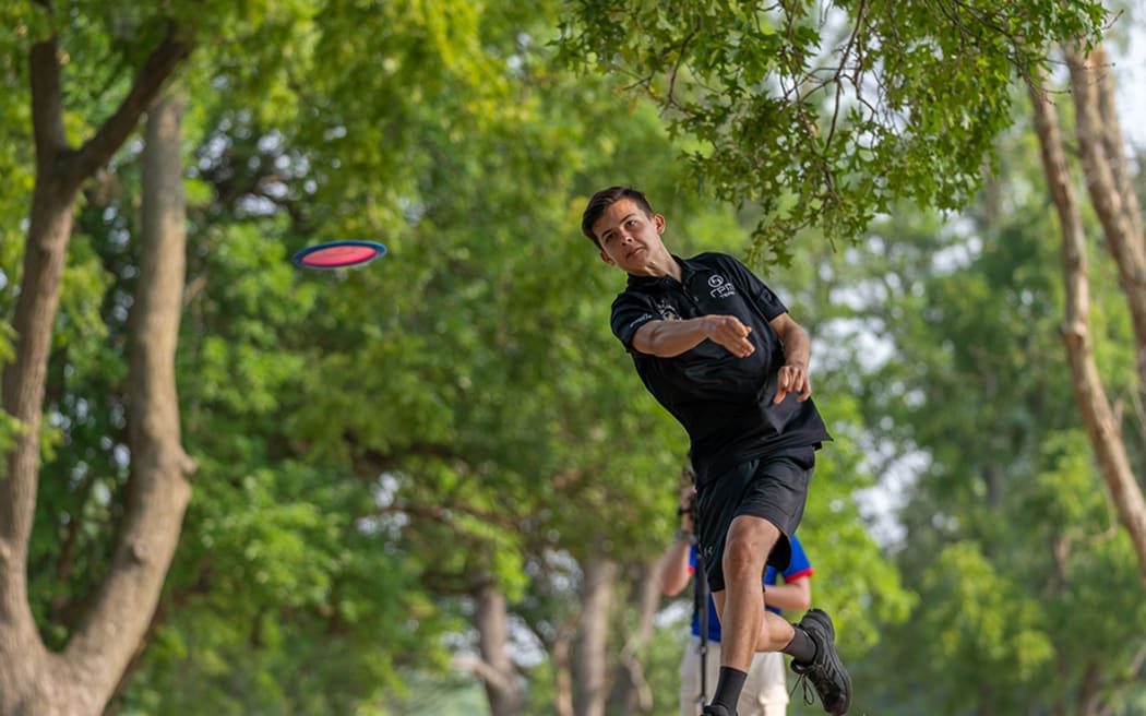 Levi throws a frisbee in a park. He wears a black polo shirt.