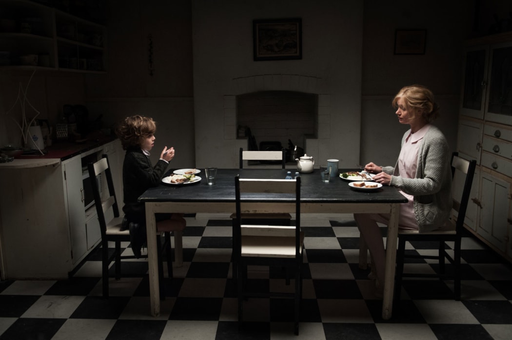 A mother and son sit at opposite ends of a dining table, in a dingy kitchen