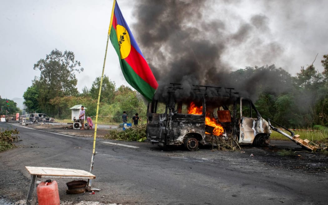 This photograph shows a Kanak flag waving next to a burning vehicle at an independantist roadblock at La Tamoa, in the commune of Paita, France's Pacific territory of New Caledonia on May 19, 2024. French forces smashed through about 60 road blocks to clear the way from conflict-stricken New Caledonia's capital to the airport but have still not reopened the route, a top government official said on May 19, 2024. (Photo by Delphine Mayeur / AFP)