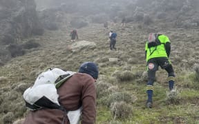 People in outdoors gear and wearing backpacks climb up a steep tussocked mountain side towards the foggy peaks,