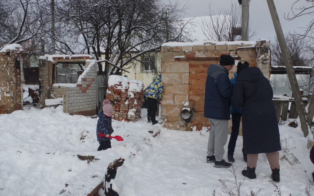Two children and three adults stand among what remains of a brick house hit by a missile strike in Ukraine. It is winter, so they are wearing big jackets and there is snow on the ground.