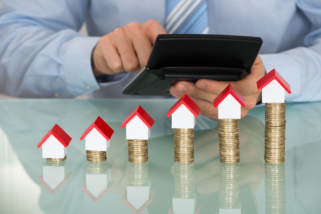 A man with a calculator inspects a line of houses stacked on top of gold coins.