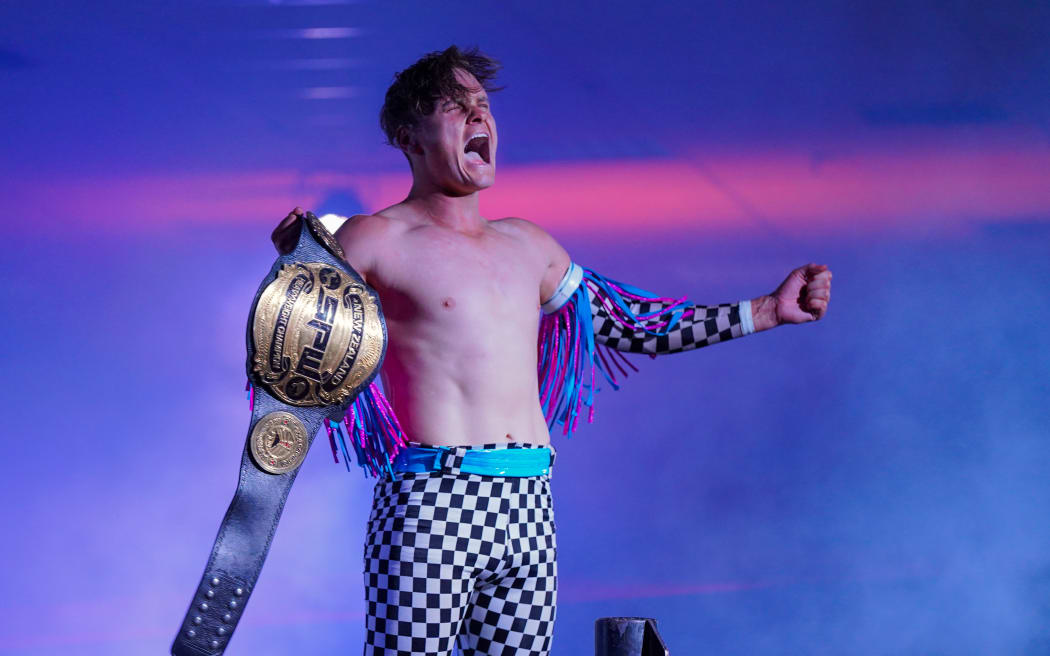 A shirtless man wearing checked leotard bottoms, holds up a gold wrestling championship belt, while screaming in joy.