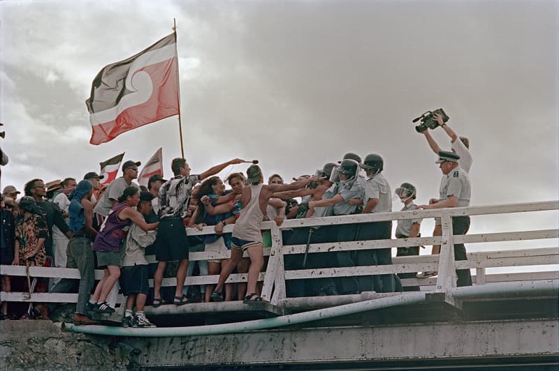 An emotive photograph capturing a confrontation between Māori protesters and police on the Waitangi bridge. A large Tino Rangatiratanga flag flies prominently on the left as a dense crowd of protesters clashes with a line of police officers in riot gear. On the right, a cameraman films the scene from behind the police line under an overcast sky.