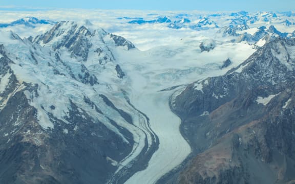 An aerial photo of a glacier snaking between tall snow-capped mountains, strewn with patches of low cloud beneath a blue sky.