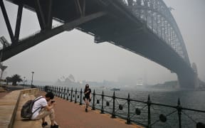 Tourists wearing masks take photos under the Sydney Harbour Bridge enveloped in haze caused by nearby bushfires, in Sydney on December 10, 2019.