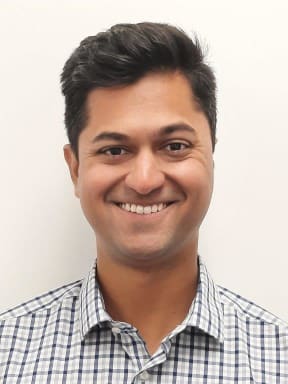 A headshot of a man wearing a white and blue checkered shirt. He is smiling directly at the camera.