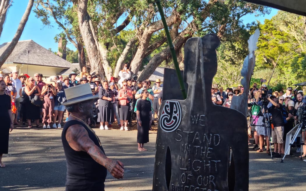 Three wāhine Māori wearing black stand in a line, their voices interlock in karanga. Tame Iti unveils a sculpture, gifting it to Waitangi and Ngāpuhi.