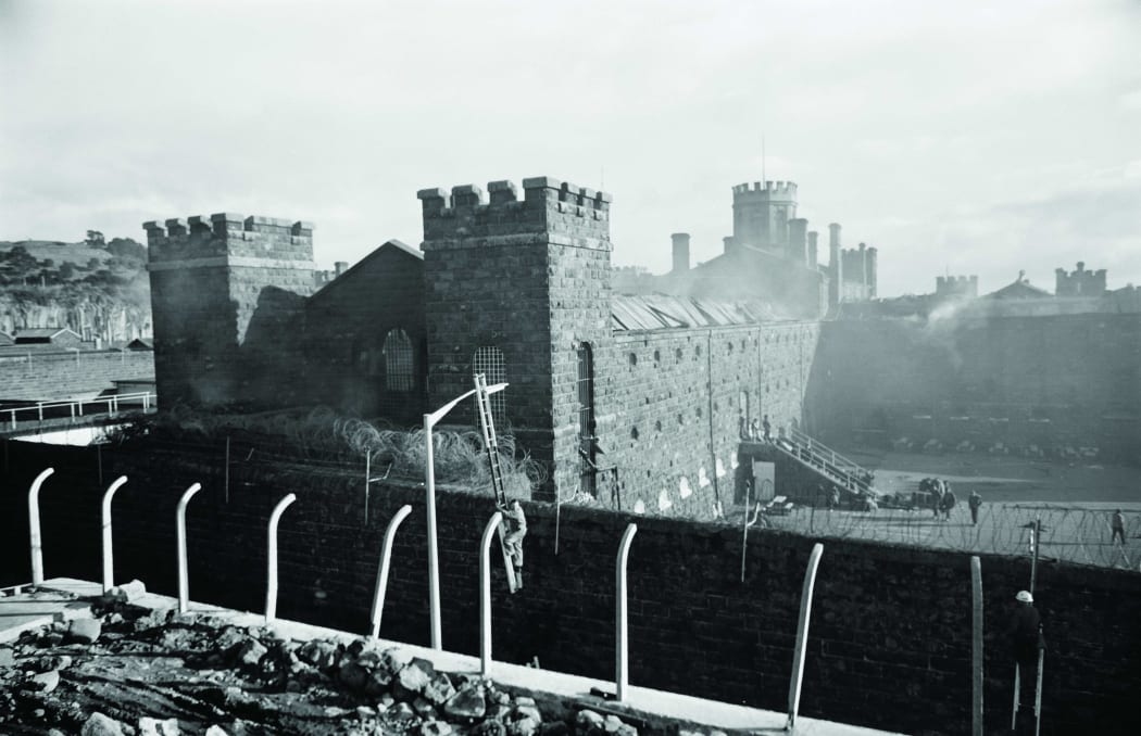 Mount Eden Prison on fire during rioting in 1965. In the foreground, Auckland Electric Power Board staff erect floodlights.