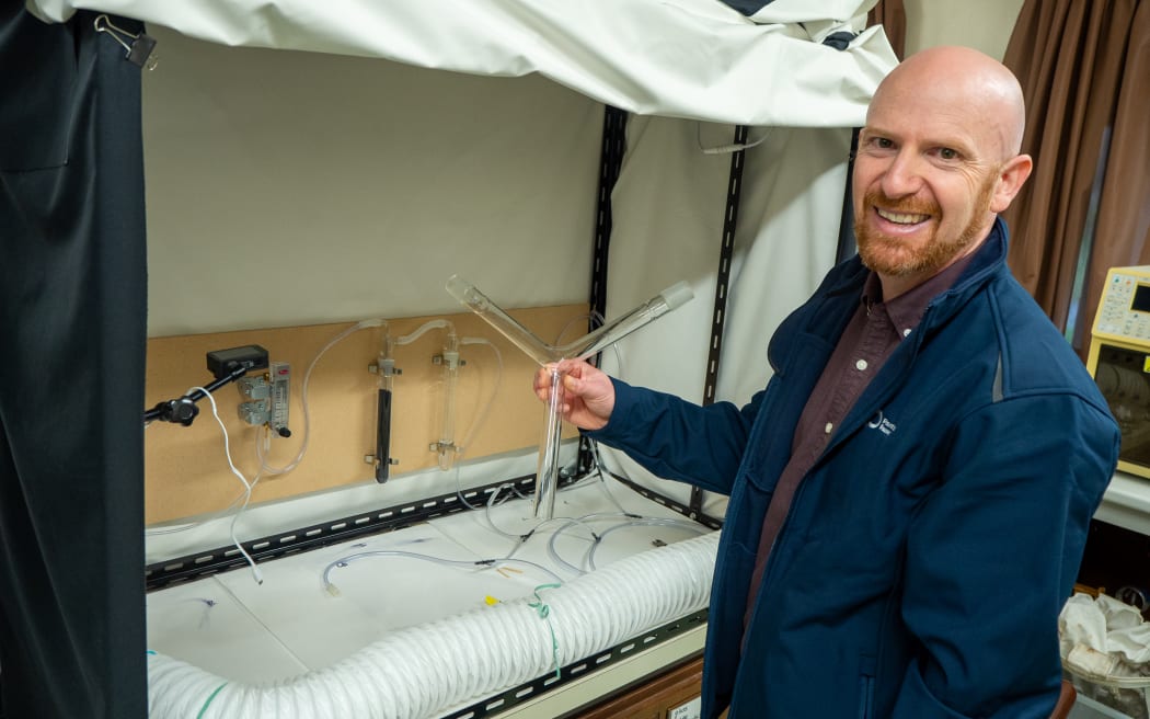 A man in a navy lab coat stands in front of a fumehoods draped with blackout sheets, holding up a glass y-shaped tube. He is smiling at the camera.