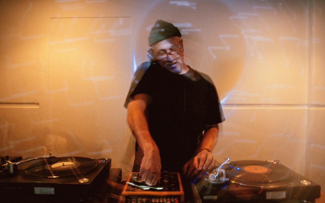 A man in a green hat and black tee-shirt DJs; he has two turntables and a sound desk set up.