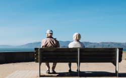 Two elderly people sit on a bench looking at a view of sea and hills.