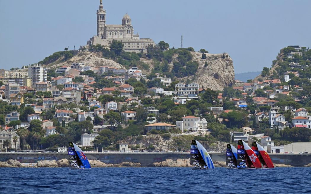 Competitors sail in front of Notre-Dame de la Garde Basilica as they compete in Race 1 of the men’s 49er skiff event during the Paris 2024 Olympic Games sailing competition at the Roucas-Blanc Marina in Marseille on July 28, 2024. (Photo by NICOLAS TUCAT / AFP)