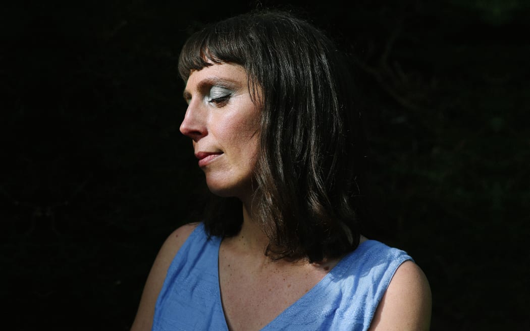 Amiria Grenell in blue dress, eyes closed in front of a black background.