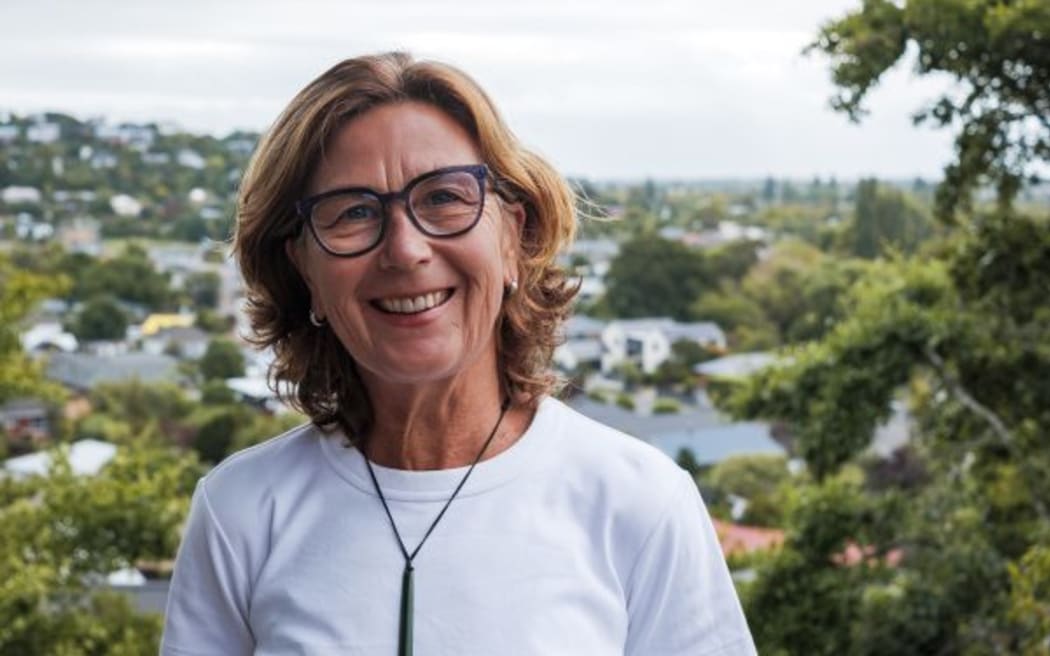 Rebecca Macfie, a middle-aged woman wearing a white top and black glasses, smiles at the camera. She stands outside in front of trees.